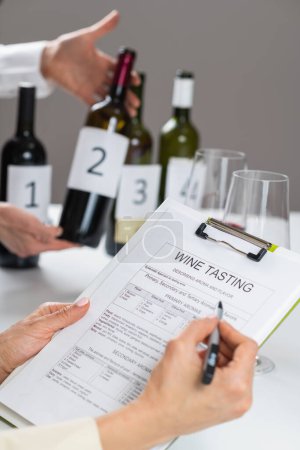 Photo for Filling out a wine tasting form. Wine tasting guest evaluates the characteristics of a red wine and fills out a wine tasting form to record observations. - Royalty Free Image