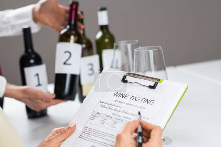 Foto de Rellenar un formulario de degustación de vinos. Los huéspedes de la cata de vinos evalúan las características de un vino tinto y llenan un formulario de cata de vinos para registrar las observaciones.. - Imagen libre de derechos