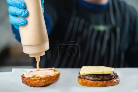 Téléchargez les photos : Cuisinier professionnel ajoutant des condiments sur un sandwich burger. Mains dans les gants en ajoutant de la sauce crémeuse sur un pain tranché. - en image libre de droit