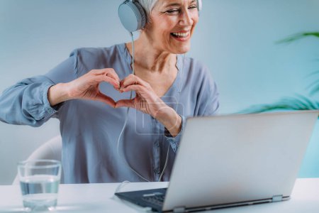 Photo for Senior woman using a laptop, having a video call with loved ones and sending love, heart hand sign. - Royalty Free Image