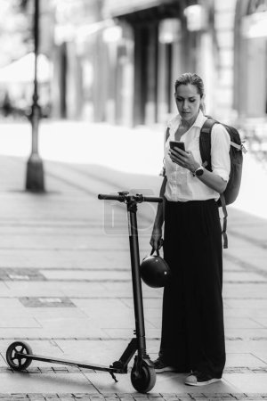 Téléchargez les photos : Femme d'affaires debout et utilisant son smartphone avec scooter électrique à côté d'elle dans la ville. - en image libre de droit