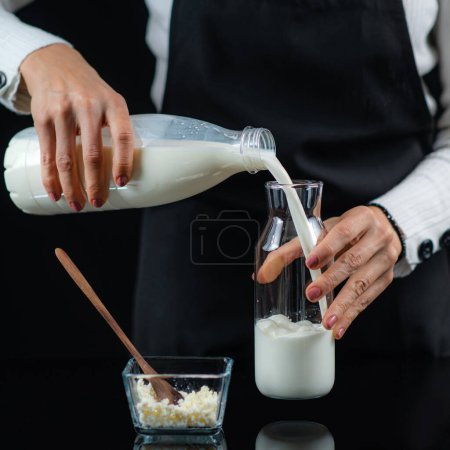 Photo for Woman Pouring milk into a glass bottle with kefir culture grains, making homemade kefir. Black background. - Royalty Free Image