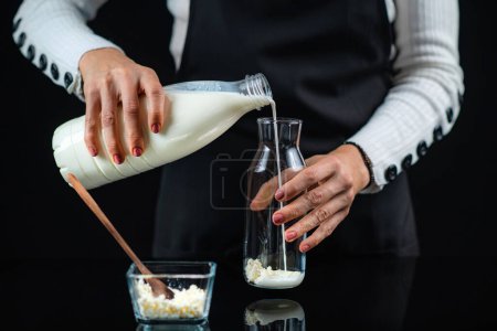 Téléchargez les photos : Femme Verser du lait dans une bouteille en verre avec des grains de culture de kéfir, ce qui rend le kéfir fait maison. Fond noir. - en image libre de droit