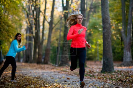 Photo for Woman Exercising in Public Park with Personal Trainer in the Fall. Trainer Cheering and Woman Running. - Royalty Free Image