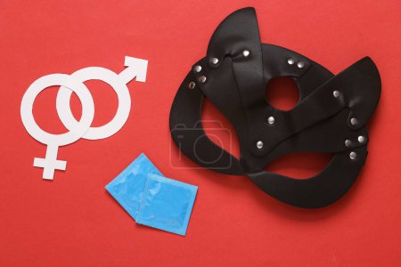 Téléchargez les photos : Masque de chat en cuir BDSM avec paquets de préservatifs et de symboles de genre sur fond rouge. Jeux de rôle jeux sexuels - en image libre de droit