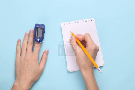 Foto de Dedo de mano con oxímetro de pulso y datos de escritura a mano en un cuaderno sobre fondo pastel azul. Medir la saturación de hemoglobina con oxígeno en la sangre capilar arterial. - Imagen libre de derechos