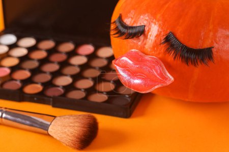 Foto de Calabaza con pestañas y labios falsos, paleta de sombras de ojos y pincel de maquillaje sobre fondo naranja. Concepto de belleza mineral, diseño creativo - Imagen libre de derechos