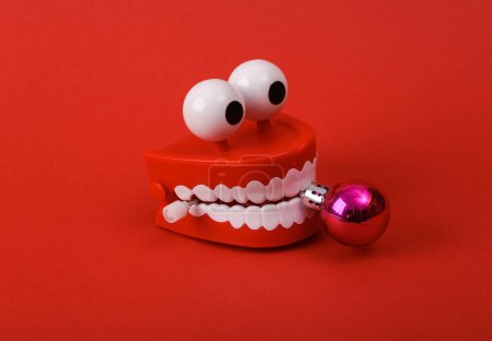 Photo for Funny toy clockwork jumping teeth with eyes holding a christmas ball on red background. - Royalty Free Image