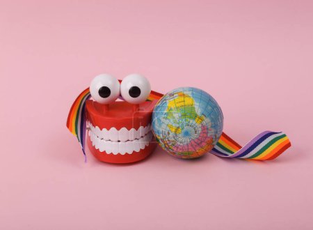 Photo for International day against homophobia. Clockwork jaw with eyes wrapped with rainbow ribbon and globe on pink background - Royalty Free Image