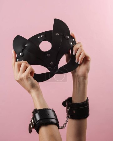 Foto de Manos de mujer en esposas de cuero con máscara de gato negro sobre fondo rosa. BDSM accesorios, dominación, juegos sexuales - Imagen libre de derechos