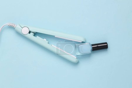 Photo for Mini hair straightener with nail polish bottle on a blue background. Pastel color. Top view - Royalty Free Image