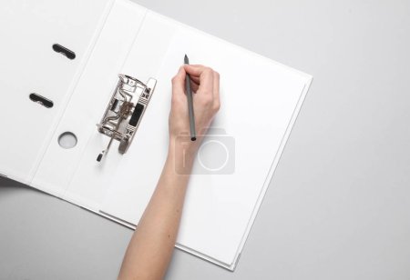 Téléchargez les photos : Femme écrit à la main sur une feuille de document dans un dossier de reliure ouvert sur fond gris - en image libre de droit