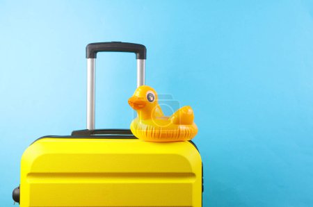 Yellow plastic travel suitcase with inflatable duck on blue background. Summer rest, tourism, vacation, relocation concept