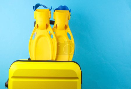 Yellow plastic travel suitcase with diving fins on blue background. Summer rest, beach vacation,