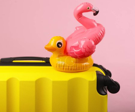 Yellow plastic travel suitcase with inflatable flamingo and duck on pink background. Summer rest, tourism, vacation, relocation concept