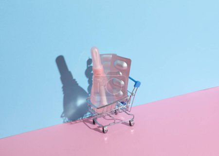 Mini carrito de supermercado con enema vaginal, pastillas sobre fondo rosa azul. Mujeres Salud