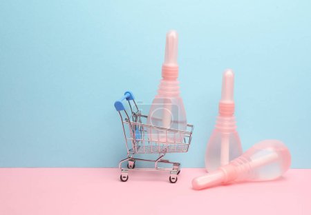 Mini carrito de supermercado con enemas vaginales sobre fondo rosa azulado. Mujeres Salud