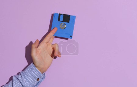 Téléchargez les photos : Main d'homme en chemise tenant une disquette bleue sur fond pastel violet avec ombre - en image libre de droit