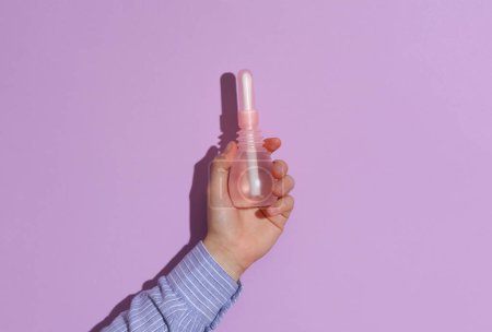 Man's hand in shirt holding Vaginal enema on purple pastel background with shadow.