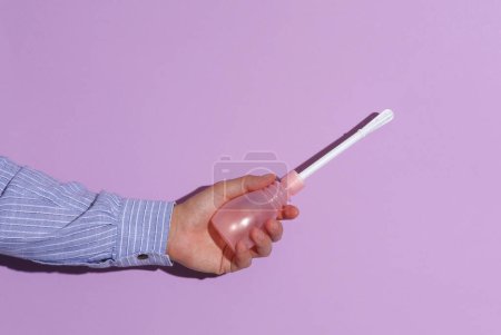 Man's hand in shirt holding Vaginal enema on purple pastel background with shadow.
