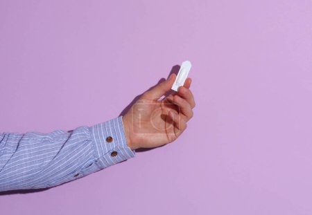 Man's hand in shirt holding rectal suppository for hemorrhoids on purple pastel background with shadow.