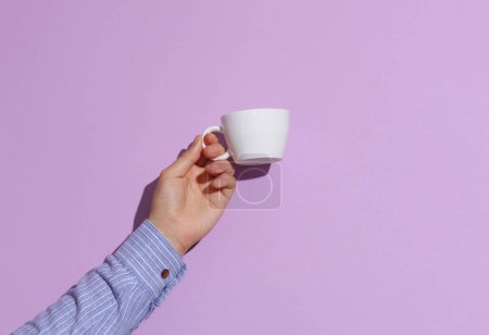 Man's hand in shirt holding white ceramic cup on purple pastel background with shadow.