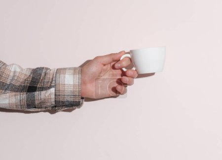 Man's hand in shirt holding ceramic cup on beige pastel background with shadow