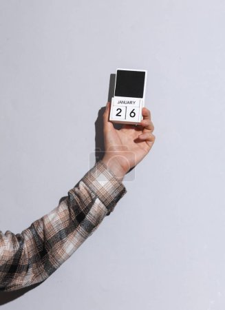 Man's hand in shirt holds wooden block calendar organizer with date january 26 on gray background with shadow