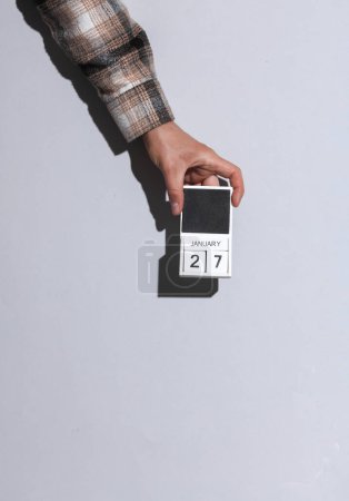 Man's hand in shirt holds wooden block calendar organizer with date january 27 on gray background with shadow
