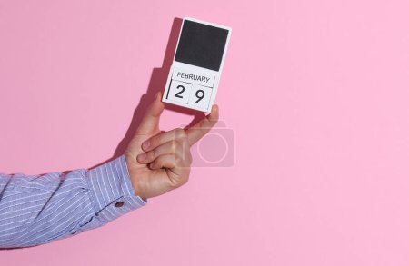 Man's hand in shirt holds wooden block calendar organizer with date February 29 on pink background with shadow