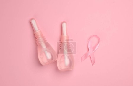 Two vaginal enemas and a pink awareness ribbon on a pink background. Women's health