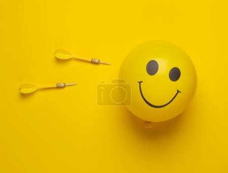 Téléchargez les photos : Ballon avec visage souriant et fléchettes sur fond jaune. Mise en page créative. Minimalisme - en image libre de droit