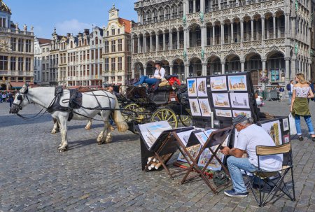 Photo for Brussels, Beigium - September 4, 2018: Paintings and carriages waiting for tourists in the Grand place (Grote Markt) square with the House of the King (City Museum) in the background - Royalty Free Image