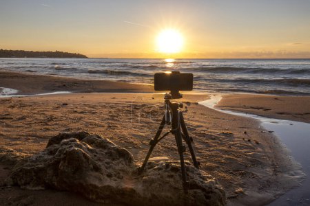 Téléchargez les photos : Un téléphone sur un trépied debout sur la plage tirant au lever du soleil - en image libre de droit