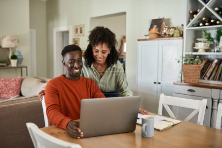 Téléchargez les photos : Jeune couple multiracial souriant tout en allant sur les factures et faire des opérations bancaires en ligne à leur table de salle à manger à la maison - en image libre de droit