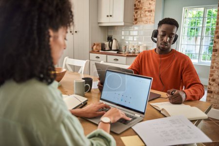 Photo for Young African man wearing headphones working remotely online from home with his wife using a laptop in their kitchen - Royalty Free Image