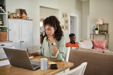 Foto de Mujer africana joven con un auricular que trabaja desde casa en un ordenador portátil con su marido sentado en un sofá en el fondo - Imagen libre de derechos