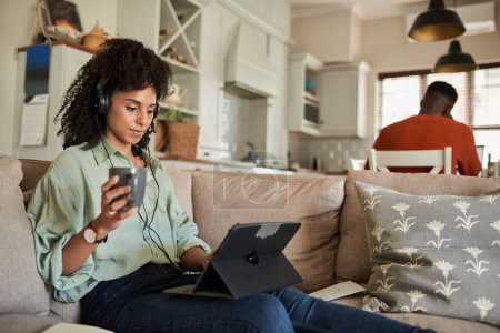 Téléchargez les photos : Young African woman wearing headphones and using a tablet on a sofa with her husband working at a kitchen table behind her - en image libre de droit