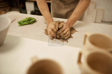Foto de Primer plano de un ceramista masculino con un delantal que trabaja con una pieza húmeda de arcilla sobre una mesa en un estudio de cerámica - Imagen libre de derechos