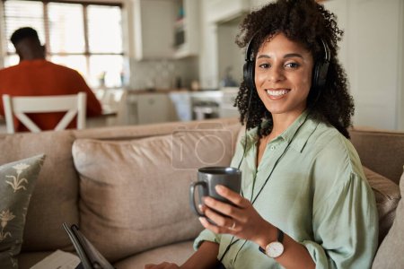 Photo for Smiling young African woman drinking coffee while wearing headphones and using a tablet on a sofa with her husband behind her - Royalty Free Image