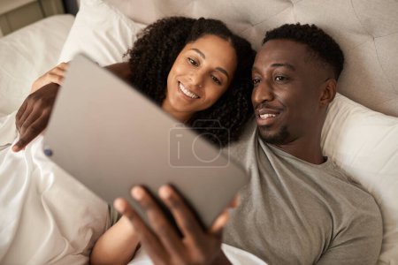 Téléchargez les photos : Souriant jeune couple multiracial utilisant une tablette numérique ensemble tout en étant couché dans leur lit à la maison le matin - en image libre de droit