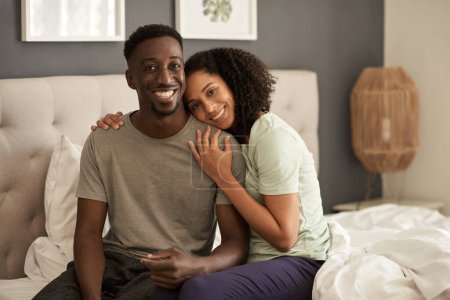 Téléchargez les photos : Portrait d'un jeune couple multiracial souriant assis ensemble sur leur lit à la maison après s'être réveillé le matin - en image libre de droit