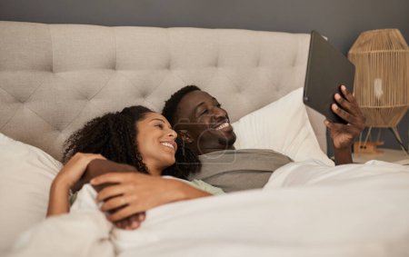Téléchargez les photos : Souriant jeune couple multiracial à l'aide d'une tablette numérique tout en étant couché ensemble dans leur lit à la maison le matin - en image libre de droit