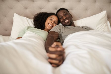 Photo for Portrait of an affectionate young multiracial couple smiling and holding hands together while lying in bed in the morning - Royalty Free Image