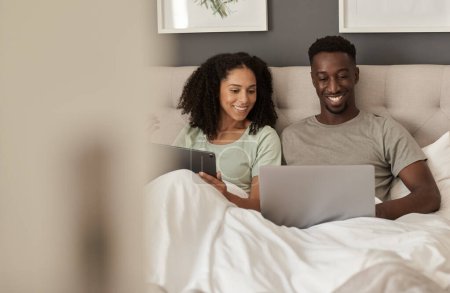 Photo for Smiling young multiracial couple sitting up in their bed at home in the morning and using a laptop and digital tablet - Royalty Free Image
