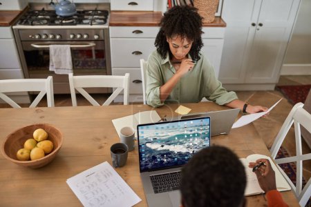 Foto de Vista de ángulo alto de una joven pareja multirracial sentada en una mesa de cocina y trabajando remotamente desde casa en computadoras portátiles - Imagen libre de derechos