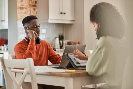 Téléchargez les photos : Jeune couple multiracial assis à une table de cuisine et travaillant à distance de la maison sur les ordinateurs portables et les téléphones - en image libre de droit