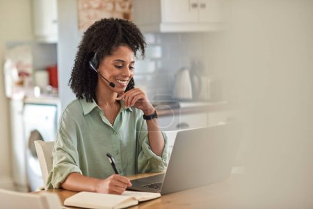 Photo for Smiling young African female entrepreneur wearing a headset talking during a video call on a laptop while working from home - Royalty Free Image