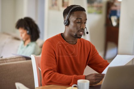 Téléchargez les photos : Jeune homme africain portant un casque de travail de la maison sur un ordinateur portable avec sa femme assise sur un canapé en arrière-plan - en image libre de droit