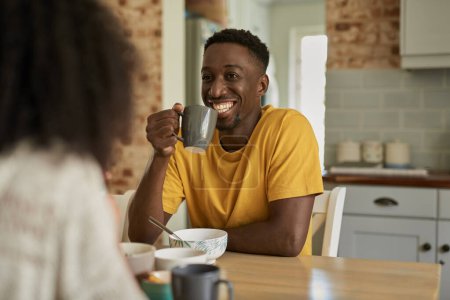 Photo for Smiling young African man drinking coffee and talking with his wife over breakfast at a kitchen table in the morning - Royalty Free Image